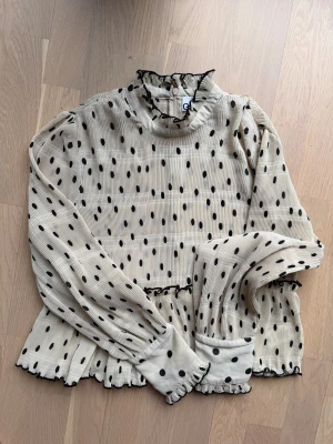 Prickig volangblus från Ganni - Supersöt beige blus från Ganni med svarta prickar, volangkrage och volangdetaljer vid ärmslut och nederkant. Blusen har lång ärm, dragkedja bak och är gjord i ett lätt, plisserat tyg som ger en luftig känsla. Perfekt för dig som gillar trendiga detaljer och lekfull stil.
