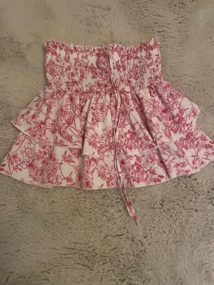 Blommig volangkjol från SHEIN - Supersöt kort kjol från SHEIN med rosa blommönster på vit botten. Kjolen har smock i midjan, volanger och en dekorativ knytning framtill. Perfekt för dig som gillar en feminin och trendig stil.