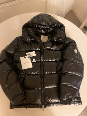  Moncler Maya - Säljer en svart  Moncler Maya med glansig finish och huva. Jackan har Moncler-logga på ärmen, dragkedja framtill och quiltad design. Perfekt för dig som vill ha en snygg och varm jacka med street-vibe. Fylld med dun och har coola detaljer som märkestaggar och justerbara muddar, NFC Scan.❄️skriv för mer frågor 🤝🏼⛷️