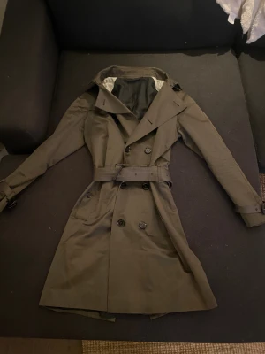 Olivgrön trenchcoat från Burberry - Snygg olivgrön trenchcoat storlek 48 vilket motsvarar Medium,  från Burberry med klassisk dubbelknäppning, bälte i midjan och spännen vid ärmsluten. Jackan har svart, glansigt foder med Burberrys logga och detaljer i svart. Perfekt för dig som vill ha en stilren och tidlös look.