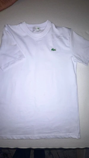 Vit Lacoste t-shirt med logga - Vit t-shirt från Lacoste i klassisk modell med rund hals och korta ärmar. På bröstet sitter den ikoniska gröna krokodilloggan broderad. Tillverkad i mjuk bomull för en skön känsla och clean look.