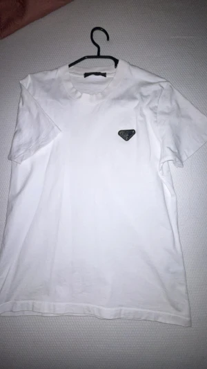 Vit t-shirt med svart logga - Snygg vit t-shirt från Prada med klassisk passform och korta ärmar. T-shirten har en svart, triangelformad logga på bröstet och är tillverkad i mjuk bomull. Perfekt för en clean och stilren look.