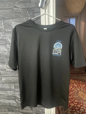 Svart Casa Blanca t-shirt med tryck - Svart t-shirt från Casa Blanca med färgstarkt tryck av palmer och stadsmotiv både fram och bak. Klassisk rund hals och korta ärmar. Snygg blå text och detaljer som ger en unik vibe. Perfekt för dig som gillar streetwear med twist.