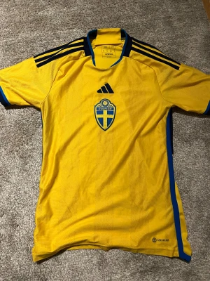 Svenska landslagets gul Adidas t-shirt - Gul t-shirt från Adidas med Sveriges landslagsemblem på bröstet och blå detaljer längs sidorna och axlarna. Klassisk sportdesign med korta ärmar, rund hals och ventilerande material. Perfekt för dig som älskar fotboll och vill representera Sverige. Från 2020