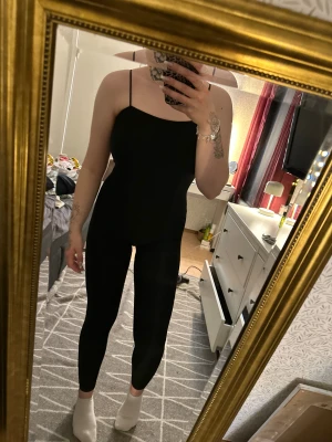 Svart byxdress med smala axelband - Svart, stilren byxdress med smala axelband och figurnära passform. Materialet är stretchigt och mjukt, vilket gör den riktigt bekväm. Byxdressen har raka ben och en enkel, minimalistisk design som är lätt att matcha med sneakers eller boots.