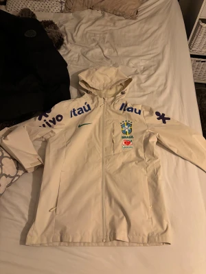 Vit Brasil Nike vindjacka M - Säljer en vit vindjacka från Nike med Brasil-logga och sponsormärken. Jackan har huva, hel dragkedja och meshfoder. Perfekt för dig som gillar fotboll och vill ha en sportig stil. Snygga blå och gröna detaljer på bröstet och ärmarna.