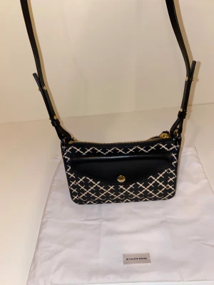 By Malene Birger axelväska - Elegant axelväska från By Malene Birger i märkets ikoniska monogrammönster. Väskan har en smidig storlek som passar vardag såväl som fest, med ett yttre fack med knäppning samt justerbar axelrem i svart läderlook. Levereras med original dustbag.