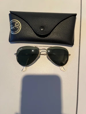 Ray-Ban Aviator solglasögon guld - Klassiska Ray-Ban Aviator solglasögon med guldfärgad metallbåge och mörkgröna glas. Kommer med svart Ray-Ban fodral i läderimitation och putsduk. Perfekt accessoar för soliga dagar och tidlös stil.