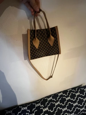 Brun och svart handväska med mönster - Snygg handväska i brun och svart med rutigt mönster och stilrena detaljer. Väskan har både handtag och axelrem, vilket gör den enkel att bära på olika sätt. Tillverkad i skinnimitation med klassisk form och fina sömmar. Perfekt för dig som gillar trendiga accessoarer.