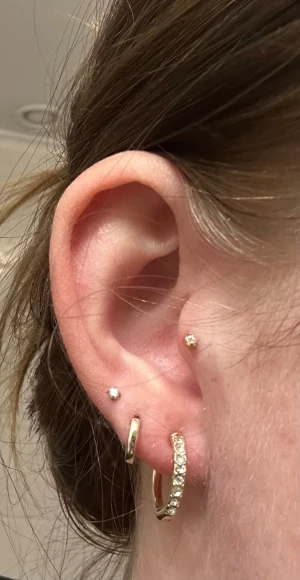 Guldiga örhängen med stenar från Glitter - Säljer ett par snygga guldiga hoops från Glitter. Ena örhänget är dekorerat med glittrande stenar längs framsidan, medan det andra är helt slätt och stilrent. Perfekt för dig som gillar att mixa och matcha smycken med lite extra bling.