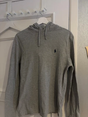 Grå hoodie från Polo Ralph Lauren - Snygg grå hoodie från Polo Ralph Lauren med klassisk broderad logga på bröstet. Tröjan har huva med snörning och är tillverkad i mjuk bomull. Perfekt för chill dagar och enkel att matcha med jeans eller joggers. Är i storlek 170 inte S 
