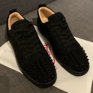 Christian Louboutin svarta sneakers med nitar - Svarta sneakers från Christian Louboutin i mocka med svarta nitar på tån och klassisk röd sula. Skorna har snörning och en stilren, edgy look som sticker ut. Perfekta för dig som vill ha något extra coolt och exklusivt i garderoben.