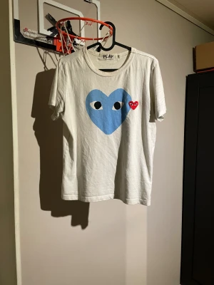 Vit Comme des Garçons Play t-shirt - Säljer en vit t-shirt från Comme des Garçons Play med ikoniskt hjärtmotiv på bröstet, ett stort blått hjärta med ögon och ett mindre rött hjärta. T-shirten har rund hals och korta ärmar, tillverkad i mjuk bomull. Perfekt för dig som gillar streetwear och coola prints. storlek S men den sitter litet så det är mer som XS