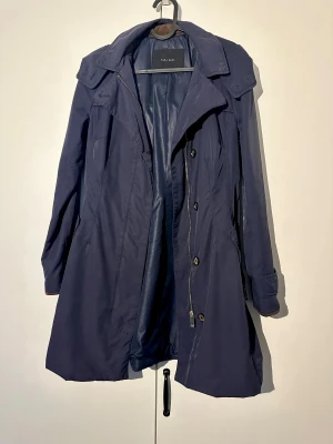 Zara Basic marinblå trenchcoat, storlek S - Mörkblå elegant trenchcoat från Zara Basic med avtagbar huva. Jackan har dragkedja och knappar framtill samt fickor med dragkedja. Observera att det finns mindre defekter på plagget.