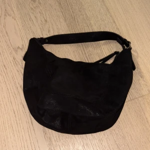 Svart axelväska i mocka - Snygg svart axelväska i mjuk mocka med rundad form och bred axelrem. Väskan har dekorativa tofsar vid dragkedjan och en stilren look som passar till många outfits. Perfekt storlek för att få plats med allt du behöver.