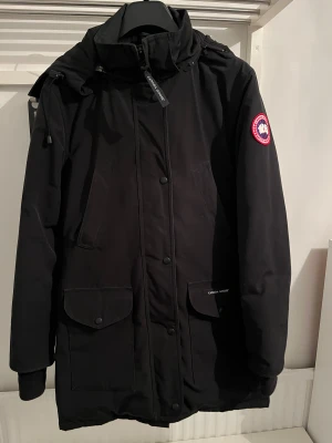 Svart parkas från Canada Goose - Svart parkas från Canada Goose med huva, dragkedja och knappar framtill. Jackan har två stora fickor med lock och logotyp på ärmen. Perfekt för kalla dagar med sin långa passform och snygga, stilrena look.
