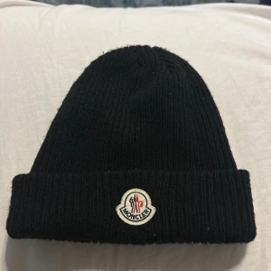 Svart Moncler mössa i ull - Svart ribbstickad mössa från Moncler med klassisk uppvikt kant och broderad logotyp framtill. Tillverkad i mjuk ull som håller dig varm och ger en clean look. Perfekt accessoar för kyliga dagar och enkel att matcha med olika outfits.