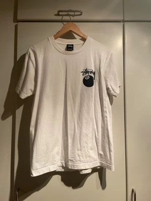 Vit Stüssy t-shirt med 8-ball print - Vit t-shirt från Stüssy med svart 8-ball tryck och logga på bröstet. Baksidan har en stor färgglad grafisk print med 8-ball, Stüssy-logga och städer som Los Angeles, New York, Paris, Tokyo och London. Baggy passform och rund hals.