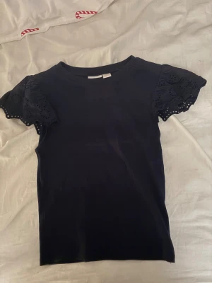 Svart blus med spetsärmar Gina Tricot - Supersöt svart blus från Gina Tricot i storlek 158/164. Toppen har korta ärmar med broderad spets och vågig kant som ger en extra detalj. Rund halsringning och mjukt bomullsmaterial gör den både bekväm och snygg. Perfekt för dig som gillar stilrena plagg med twist.