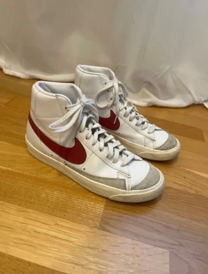 Nike Blazer 77 Mid  vit/röd sneakers - Säljer ett par klassiska Nike Blazer 77 Mid sneakers i vitt skinn med röd swoosh och detaljer. Skorna har högt skaft, snörning och grå mockadetaljer vid tån. Perfekta för dig som gillar retrostil och streetwear. Skorna är sparsamt använda.