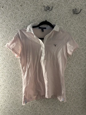 Ljusrosa pikéskjorta från GANT - Snygg ljusrosa pikéskjorta från GANT i storlek S. Skjortan har vit krage och knappslå med tre knappar framtill. Klassisk GANT-logga broderad på bröstet. Tillverkad i mjuk bomull, kortärmad och med slitsar i sidorna för extra komfort.