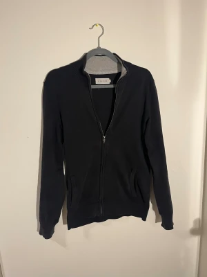 Svart kofta med dragkedja Pier One - Svart kofta från Pier One i storlek S. Tröjan har ribbad krage i grått, hel dragkedja framtill och två fickor. Materialet är mjukt och stickat, perfekt för lager på lager. Enkel och stilren design som funkar till många olika looks. Ett litet hål på armen men jag kan fixa det innan tröjan skickas!