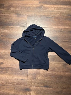 Mörkblå hoodie från Polo Ralph Lauren - Snygg mörkblå hoodie från Polo Ralph Lauren med dragkedja, känguruficka och klassisk röd broderad logga på bröstet. Tillverkad i mjuk bomullsblandning och har en stor huva för extra chill vibe. Perfekt för dig som vill ha en clean och stilren look.