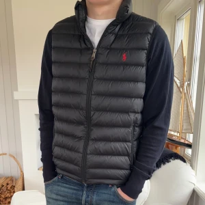 Svart pufferväst från Polo Ralph Lauren - Svart pufferväst från Polo Ralph Lauren med röd logga på bröstet. Västen har ståkrage, horisontella quiltade sektioner och dragkedja framtill. Materialet är syntet och dun, perfekt för lager-på-lager under höst och vinter. Snygg och stilren design som funkar till jeans.