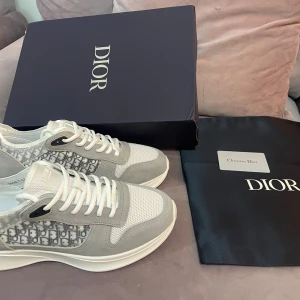 Grå och vita sneakers - Snygga sneakers i grått och vitt. Skorna har snörning, rund tå och platt sula. Ovandelen är i mesh och mocka med exklusiva detaljer och logotyp på plösen. Perfekt för dig som gillar lyxiga och trendiga sneakers.
