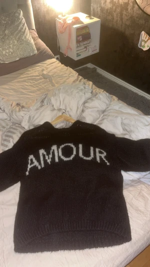 Svart stickad tröja från Noella XS/S - Svart stickad tröja från Noella i storlek XS/S med vit/ beige text 'AMOUR' framtill. Tröjan har en loose passform och långa ärmar, perfekt för dig som gillar chill och trendiga statement-plagg. Mjuk och fluffig känsla, passar till jeans eller kjol.