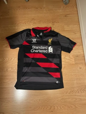 Liverpool FC Warrior fotbollströja M - Säljer en Liverpool FC fotbollströja från Warrior i svart och grått med röda diagonala ränder och detaljer. Tröjan har korta ärmar, röd krage och klubbmärke i guld på bröstet. Materialet är lätt polyester, perfekt för fotboll och sport.