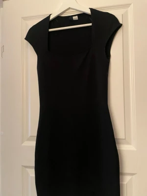 Svart kortklänning med fyrkantig ringning - En stilren svart kortklänning med fyrkantig ringning och korta ärmar. Klänningen har en figurnära passform och är enkel men elegant i designen. Perfekt för dig som gillar minimalistisk stil. Passar även mig som har xs/s