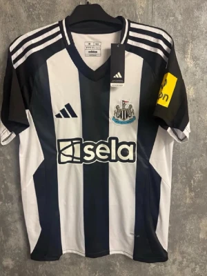 Newcastle United tröja Isak 14 Adidas - Säljer en officiell Newcastle United matchtröja från fotbollsmarknaden med Isak och nummer 14 på ryggen. Tröjan har klassiska svarta och vita ränder, Premier League-logga och sponsortryck. V-ringad hals och gula detaljer på ärmen. Perfekt för fotbollsfans! Vid snabbaffär är priset ej hugget i sten, skriv för mer bilder