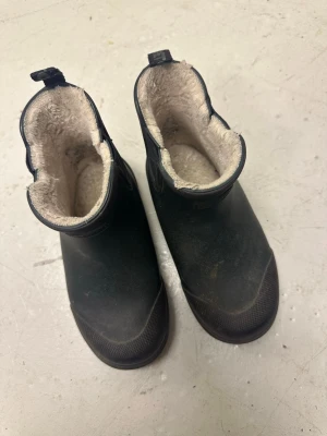 Svarta fodrade boots från Tretorn - Säljer ett par svarta boots från Tretorn med varm, beige fuskpäls på insidan. Skorna är tillverkade i slitstark gummi med grov sula för extra grepp. Praktisk ögla bak för enkel påtagning och diskret logga på sidan.