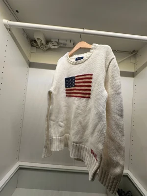 Vit stickad tröja Polo Ralph Lauren - Vit stickad tröja från Polo Ralph Lauren med amerikansk flagga broderad på bröstet och RL-broderi i rött vid nederkanten. Rund hals och ribbade muddar vid ärmslut och midja. Perfekt för dig som gillar klassisk och cool stil.   Varm och skön!     Passar för dig som är vid längden 168cm👍 Nästan aldrig använd.   Ny pris 2900kr.  Bästa dealen för dig som inte vill spendera för mycket pengar 