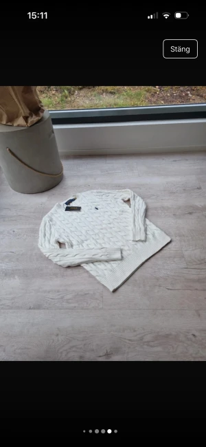 Vit kabelstickad tröja från Polo Ralph Lauren - Vit kabelstickad tröja från Polo Ralph Lauren med klassisk blå broderad logga på bröstet. Tröjan har rund hals, ribbade muddar och nederkant samt långärmad passform. Perfekt för dig som gillar preppy stil och vill ha något mjukt och snyggt till hösten.