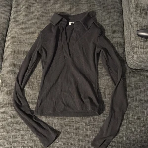 Svart ribbad långärmad topp NLY Trend - Säljer en svart ribbad långärmad topp från NLY Trend i storlek S. Toppen har en djup v-ringning och klassisk krage, vilket ger en snygg och stilren look. Perfekt att matcha med jeans eller kjol för en trendig outfit.