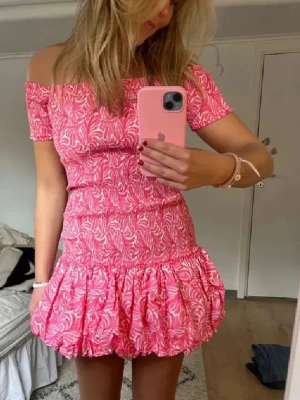 Rosa offshoulder kortklänning - Supersöt rosa kortklänning med offshoulder och volangkant. Klänningen har ett vitt bladmönster, smockad överdel och korta ärmar. Perfekt för sommarens alla tillfällen och ger en riktigt fräsch vibe❤️ Klänningen är oanvänd och har prislappen på sig.