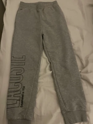 Grå mjukisbyxor från Lacoste - Grå mjukisbyxor från Lacoste med resår i midjan och muddar vid bensluten. Stort Lacoste-tryck längs ena benet och klassiska krokodilloggan vid fickan. Tillverkade i mjuk bomullsmix för maximal komfort.