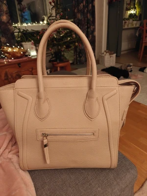 Beige handväska i skinn med dragkedja!  - Säljer en snygg beige handväska i skinn med två rejäla handtag och en praktisk dragkedja framtill. Väskan har en stilren och strukturerad design med detaljerade sömmar och gott om plats för allt du behöver. Perfekt accessoar för att lyfta din outfit. Inte äkta! 