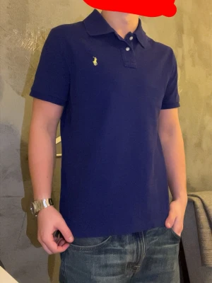 Blå pikétröja från Polo Ralph Lauren - Säljer en klassisk blå pikétröja från Polo Ralph Lauren i storlek XL. Tröjan har korta ärmar, krage och två knappar framtill. Ikoniska gula loggan broderad på bröstet. Skön passform och perfekt till jeans.