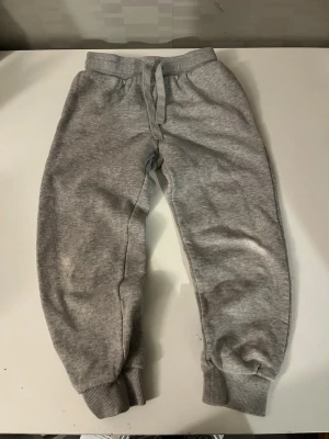 Grå mjukisbyxor från H&M med snörning - Grå mjukisbyxor från H&M i storlek 116. Byxorna har resår i midjan med snörning och muddar vid bensluten. Tillverkade i en mjuk bomulls- och polyesterblandning som känns skön mot huden. Perfekta för chill dagar eller när du vill vara bekväm.