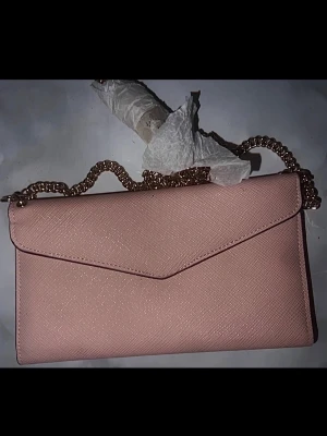 Rosa kuvertväska med guldkedja till iPhone  - Snygg rosa kuvertväska med guldfärgad kedja som axelrem. Väskan har ett stilrent, lätt glittrigt yttre och praktiska kortfack på insidan. Perfekt för att bära mobilen och det viktigaste. Enkel och elegant design.