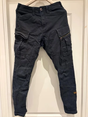 Svarta cargobyxor från G-Star RAW - Säljer ett par svarta cargobyxor från G-Star RAW i storlek 31/34. Byxorna har flera stora fickor med både dragkedja och lock, samt detaljerad sömnad och patch med logga på benet. Tillverkade i slitstark bomull och har en tapered passform.