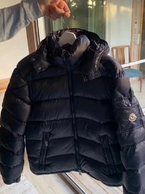  Moncler pufferjacka - Säljer en marinblå pufferjacka från Moncler med klassisk logga på ärmen och coolt brunt innerfoder med serietidningsdetalj. Jackan har hög krage, dragkedja framtill och två fickor med dragkedja. Perfekt för kalla dagar och riktigt snygg streetstil.