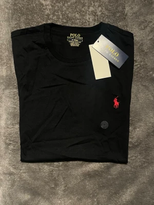 Svart t-shirt från Polo Ralph Lauren - Svart t-shirt från Polo Ralph Lauren i custom slim fit. Klassisk design med rund hals och korta ärmar. Ikoniska röda broderade loggan på bröstet. Tillverkad i mjuk bomull för skön känsla och stilren look.