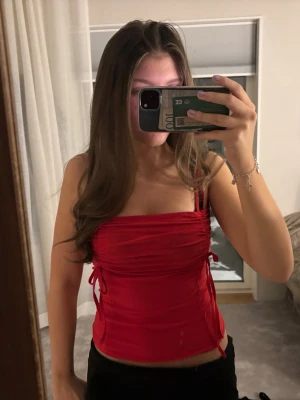 Röd croppad linnetopp med snörning - Snygg röd croppad linnetopp med smala axelband och snörningsdetaljer på sidorna. Toppen har en tight passform och är tillverkad i ett mjukt, stretchigt syntetmaterial. Perfekt för dig som vill sticka ut med en färgstark look.