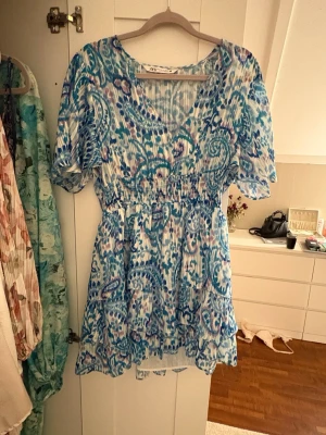 Blåvit kortklänning från Zara - Supersöt kortklänning från Zara med blått och vitt paisleymönster, v-ringning och korta ärmar. Klänningen har smock i midjan och volangdetaljer i kjolen. Perfekt för sommarens alla dagar!