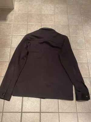 Svart overshirt  - Säljer en svart overshirt med klassisk krage och tryckknappar framtill. Två stora bröstfickor med lock och knappdetaljer ger en clean look. Perfekt lager-på-lager-plagg för dig som gillar minimalistisk stil.