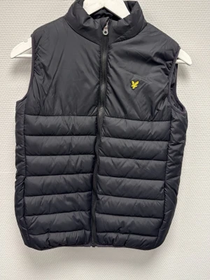 Sv trademarks dunväst från Lyle & Scott - Snygg svart dunväst från Lyle & Scott Junior med gul logga på bröstet. Västen har hög krage, quiltad design och dragkedja framtill (full zip). Perfekt att slänga över en hoodie för en chill look. Skriv för frågor!